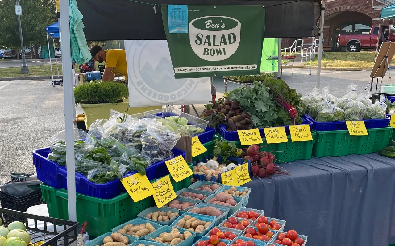 Ben's Salad Bowl produce stand at the farmers market in Lewisburg, West Virginia, with tomatoes, greens, potatoes, beets, and other produce displayed under a canopy.